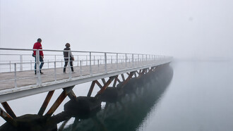 Pedestrians crossing a long wooden bridge over calm water on a misty, fog-covered morning