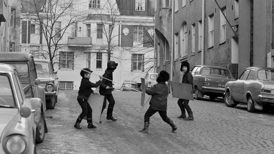 Eeva Rista, Four boys fencing with sticks and shields at the end of Meritullinkatu. Meritullinkatu 32 to the left and Meritullinkatu 33 to the right. Kristianinkatu 7 in the centre, January 1971. Courtesy of Helsinki City Museum