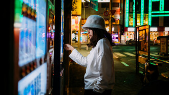 An Asian woman selects a beverage from a smart touchscreen vending machine in a brightly lit urban setting