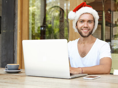 Smiling brightly, a young man in a red Santa hat browses his emails on a laptop, spreading holiday cheer