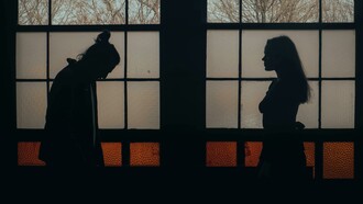A man and woman standing in a dimly lit room, surrounded by darkness and emotional tension