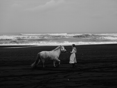 A woman walks her white horse on the beach on a grey day
