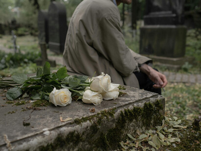 A man in a trenchcoat sitting down besides a tombstone, after leaving flowers