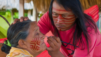 O artista Jaider Esbell pintando rosto de uma senhora de sua comunidade indígena em Roraima, Brasil