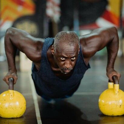 An older man demonstrates strength and determination as he performs push-ups on kettlebells in a gym setting, highlighting the importance of regular exercise and strength training