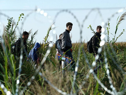 Migrants walk on an open field beside a barbed fence