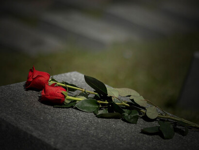 Flowers lay gently on a gravestone as a solemn burial ceremony unfolds, with mourners grieving and a funeral procession passing in the distance