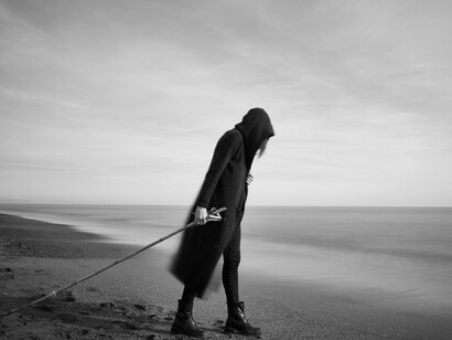A hooded figure walks by the beach holding a long pole