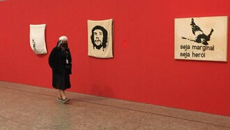 Visitante observa obras de Hélio Oiticica em exposição, entre elas a emblemática "Seja marginal seja herói". Inspirada na morte de Manuel Moreira, conhecido como "Cara de Cavalo", a criação tornou-se um manifesto artístico contra a violência e a exclusão, reafirmando a capacidade da arte de desafiar convenções — movimento que também marcou a literatura brasileira a partir da Geração de 1945. Ao transformar um episódio de violência em reflexão artística, Oiticica ajudou a redefinir os caminhos da arte contemporânea brasileira, em sintonia com a renovação da literatura e de outras formas de expressão