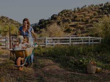 Happy mother joyfully pushing her two daughters seated inside a wheelbarrow amidst a picturesque rural European landscape, capturing the essence of family holidays in the countryside