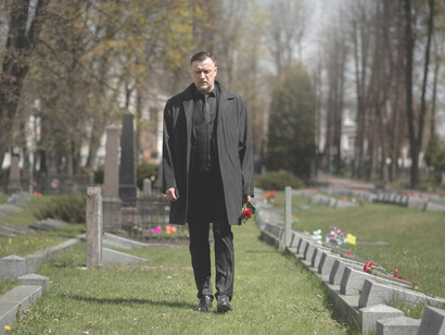 A man visiting a cemetery, mourning at a gravesite, while a funeral procession takes place nearby
