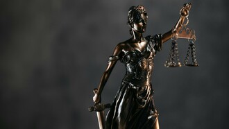 A sculpted defender of law with suspended scales, representing the fragile balance of democratic institutions
