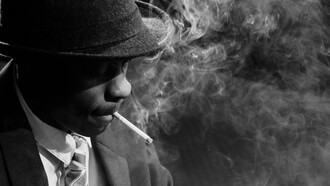 Um homem negro de terno usando um chapéu e fumando um cigarro em meio a redemoinhos de fumaça