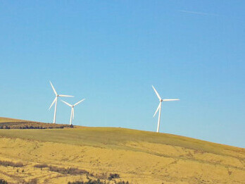Three white windmills rise proudly above the lush green fields of Cymmer, Wales, set against a vibrant blue sky, capturing the essence of sustainable energy in harmony with nature