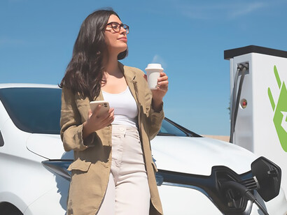 The delight of an upbeat woman relishing her coffee while eagerly awaiting her electric car to charge – a snapshot of eco-friendly travel and sustainable living that resonates with positive energy