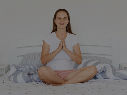 Indoor shot of a dark-haired woman with a charming smile, dressed in a white casual t-shirt and shorts, sitting in a lotus pose on a bed in a light, serene bedroom, meditating with palms together in the morning