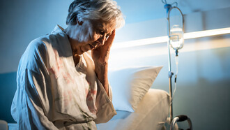 An elderly woman sits alone in a hospital room, holding her head in her hand