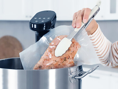 Concerning use of plastic as a woman places vacuum-sealed meat into a pot with a sous vide cooker, highlighting the detrimental impact of plastic usage in kitchen practices