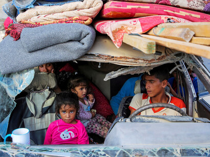 In a visibly battered car, a father and his children flee the devastation of war, carrying their remaining possessions as they search for safety