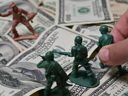 Lifesize toy soldier engaged in combat, shooting at an enemy figure amidst a backdrop of scattered dollar bills, symbolizing the intersection of play and the harsh realities of war