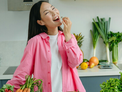 A woman in a kitchen filled with produce wearing a pink shirt smiling and eating a berry