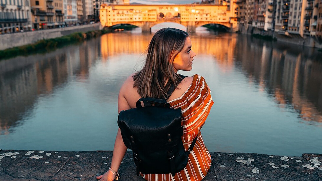 Amid the sunset's glow in Florence, Italy, a woman wears a stylish black and orange striped shirt, holding a black leather sling bag, as she stands gracefully on the gray concrete