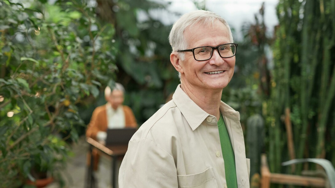 An elderly man in glasses smiling in a garden with another person in the background