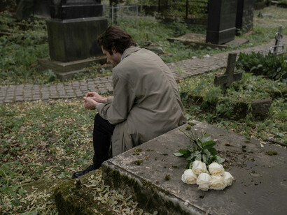 A man in a trenchcoat mourning next to a tombstone, where flowers rest