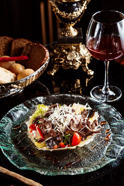Side view of a beef salad with vegetables and Parmesan cheese on a plate, paired with red wine on the table