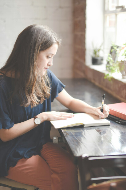 A woman at a black table, writing in a white book by the window, reflects on literary authenticity, cultural appropriation, book criticism, author identity, and creative freedom