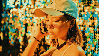 A girl wearing sunglasses and a cap, talking on the phone in front of neon lights
