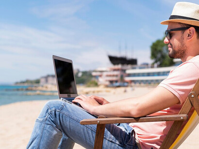 Man enjoying a drink while working on a laptop from a beach chair, illustrating flexible summer work arrangements and work-life balance