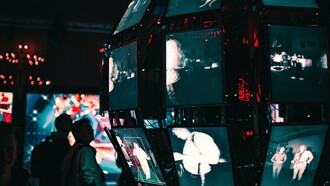 Pessoas observam dezenas de telas acesas em uma sala de baixa luminosidade. Mediante narrativas fragmentadas e imagens simultâneas, o presente parece disputar atenção a cada segundo. O excesso informativo tornou-se uma das paisagens mais características do nosso tempo. Com tanta propaganda, memória e criação, multiplicam-se as narrativas contemporâneas