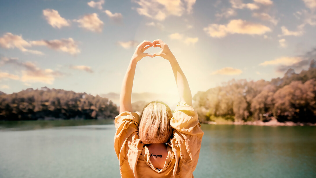 Experience a symbolic moment of self-discovery as a woman creates heart-shaped hands against a sunset backdrop in this captivating image