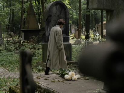 A man in a trenchcoat walking in a cemetery, visibily downtrodden