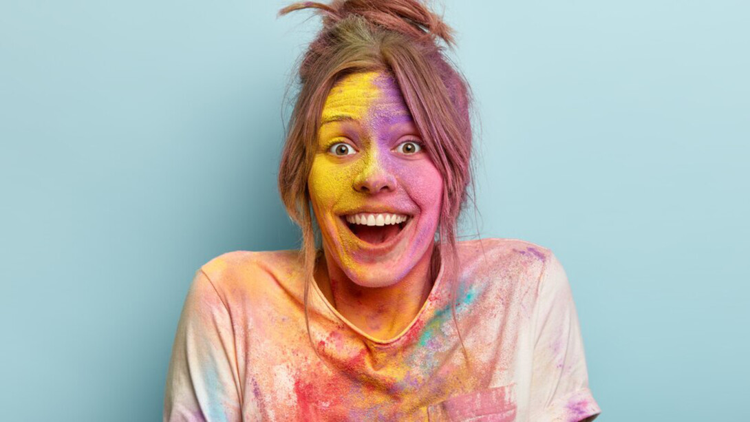 Headshot of an optimistic woman in a white t-shirt, smiling as she plays with vibrant colors, her face and clothes covered in colorful splashes