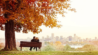 A young married couple sits on a bench, sharing a kiss as the man lovingly wraps his arm around his wife’s shoulders on a beautiful autumn day