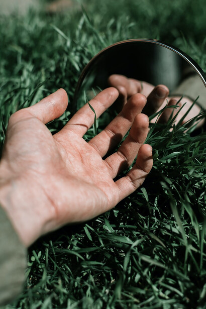 In an outdoor setting, a person holds a circular black frame, within which the reflection of the lush grass and expansive sky is captured