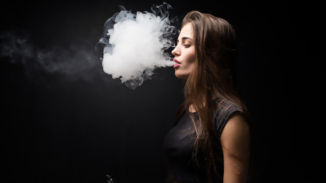 A woman vaping an e-cigarette, with smoke drifting against a black wall