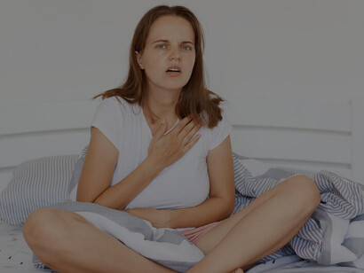 A woman in a white t-shirt, sitting cross-legged on a bed, touching her chest and looking at the camera with a concerned expression