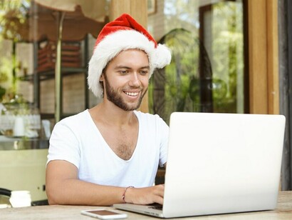 A young man wearing a festive Santa Claus hat beams at the camera as he reads his emails on his laptop