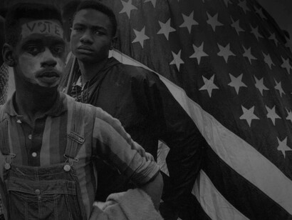 Two African-American men near the American flag, one is holding it and the other has the word "vote" painted on his forehead during election season