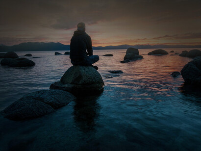 A man sitting on a rock that's on a body of war is looking contemplatively at the sunset in full meditation