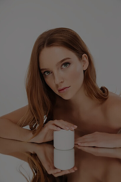Discover the allure of premium beauty products as this ginger woman, adorned with flowing locks, effortlessly displays a bottle of sumptuous body cream on a sleek mirror surface