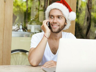 A cheerful young man in a red Santa Claus hat smiles at the camera while checking his emails on a laptop