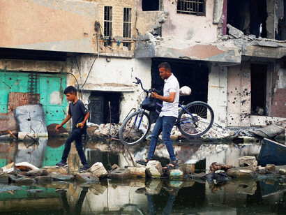 A man crosses stagnant water amidst the wreckage of the war zone, embodying the determination to move forward despite overwhelming challenges