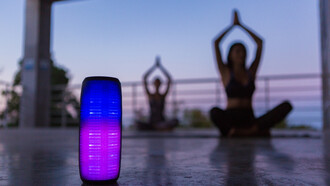 A colorful backlit wireless speaker in the foreground, as a group of friends practices yoga in the background