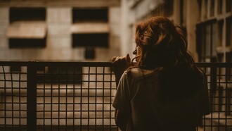 A woman stands alone on a balcony, gazing through the railing in a sepia-toned scene with a quiet and contemplative atmosphere