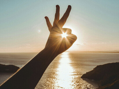 The sun passes through a circle made by a human hand, reflecting on the sea in the background