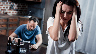 Depressed woman sitting in distress as her husband drinks alcohol in the background